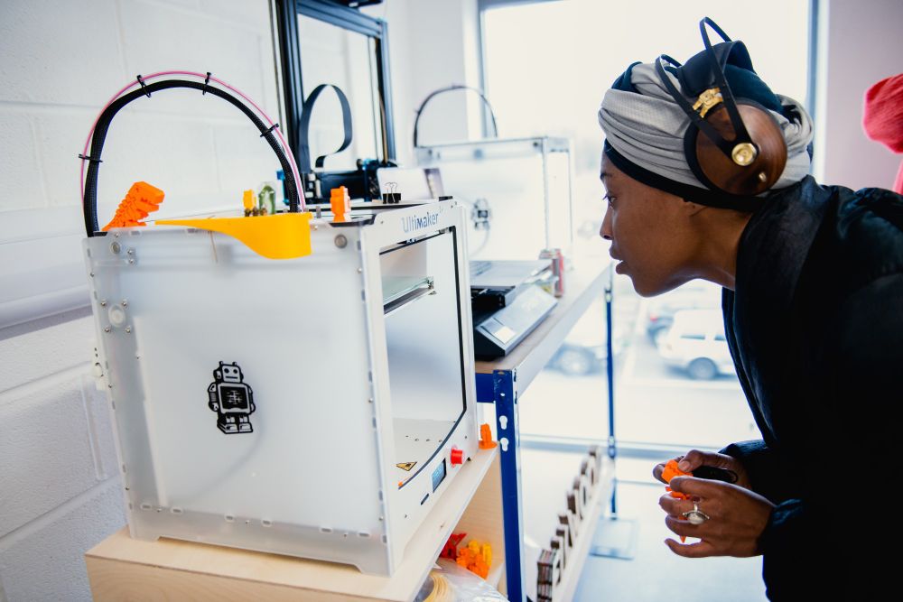 Person observing a 3D printer producing orange objects.