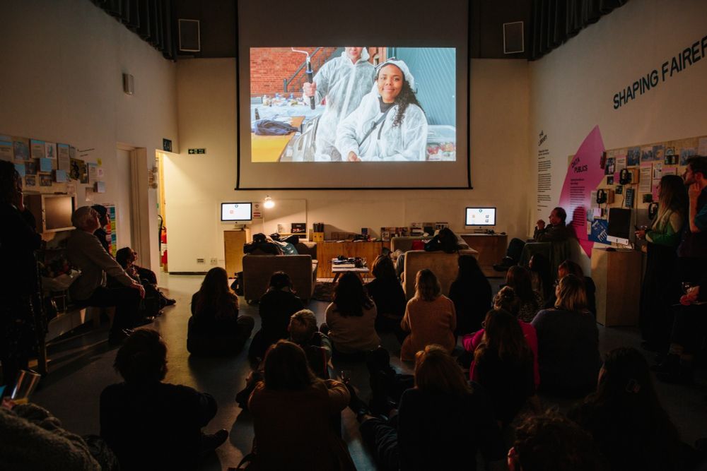 A seated audience watches a film screening in a dimly lit exhibition space, with a large projected image of two people in protective gear painting a building.