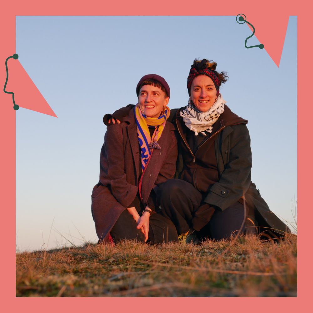Linzy and Rachael are smiling at the camera, sitting on grassy hill at golden hour, wrapped up warm in coats and scarves.