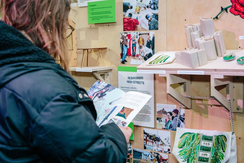 A person reads a booklet in front of an exhibition display featuring creative community projects and text about reimagining high streets.