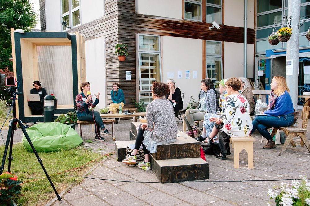 Group of people sitting in front of a modern building (KWMC). They are surrounded by a hybrid setup with cameras and microphones.