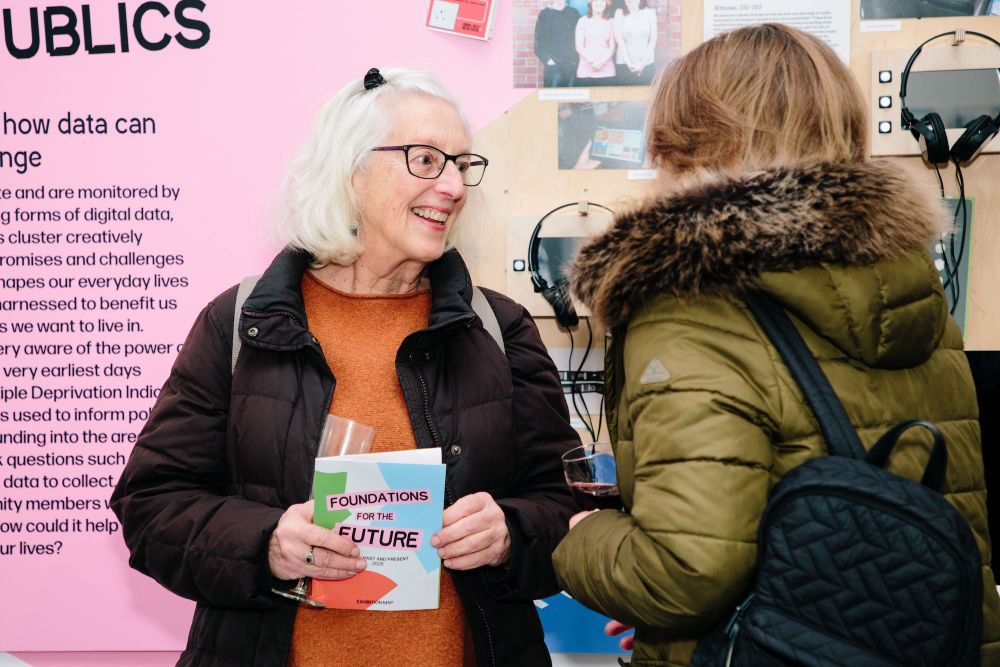 Two people chat at an exhibition, one holding a booklet titled Foundations for the Future, surrounded by displays about data and public engagement.