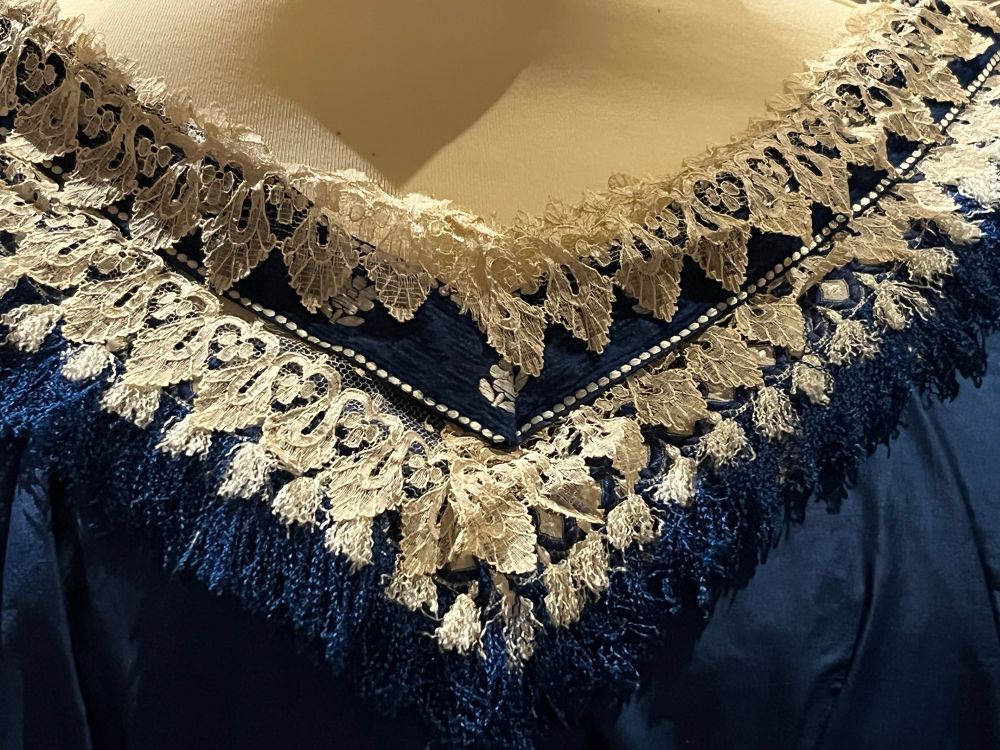 A neckline detail of an 1860s dress showing white lace and blue as well as white tassels. Photo by Robert Emond 