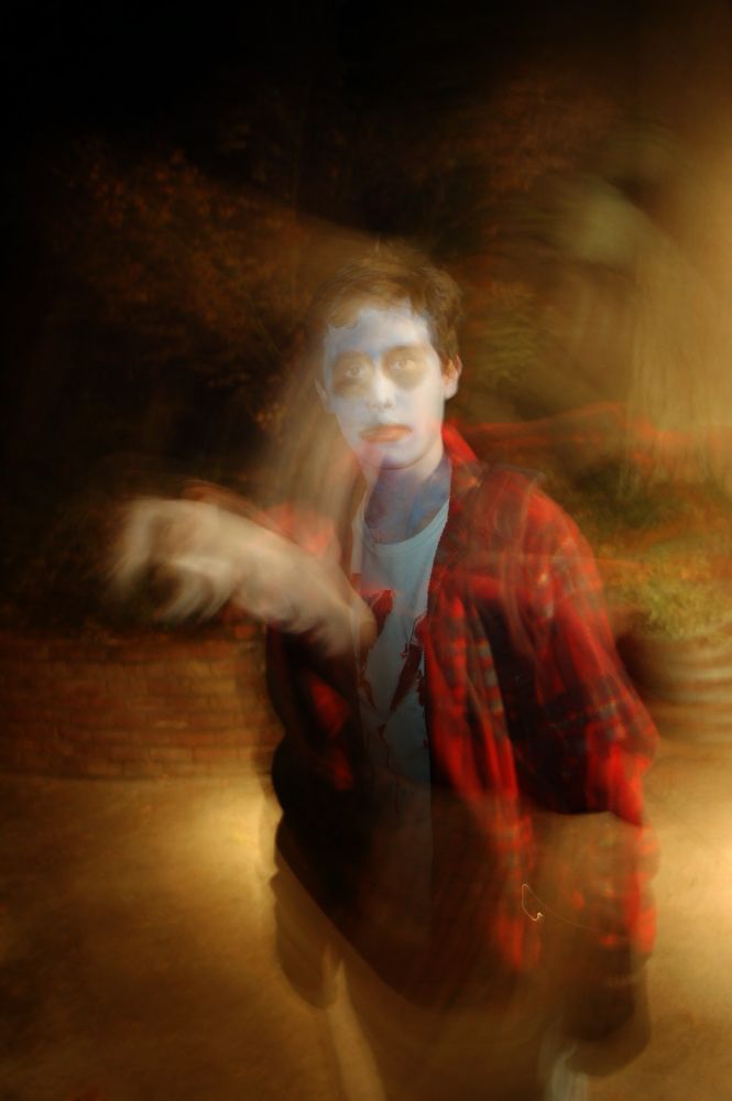 A haunting image of a young man in a red plaid shirt. There are bloody slashes on his shirt. His complexion is pale with dark eyes.  A ghostly image of his head appears to be coming out of the wound on his chest. Model, Alex Emond. Makeup and costume by Lisa Emond. Photo by Robert Emond. 
