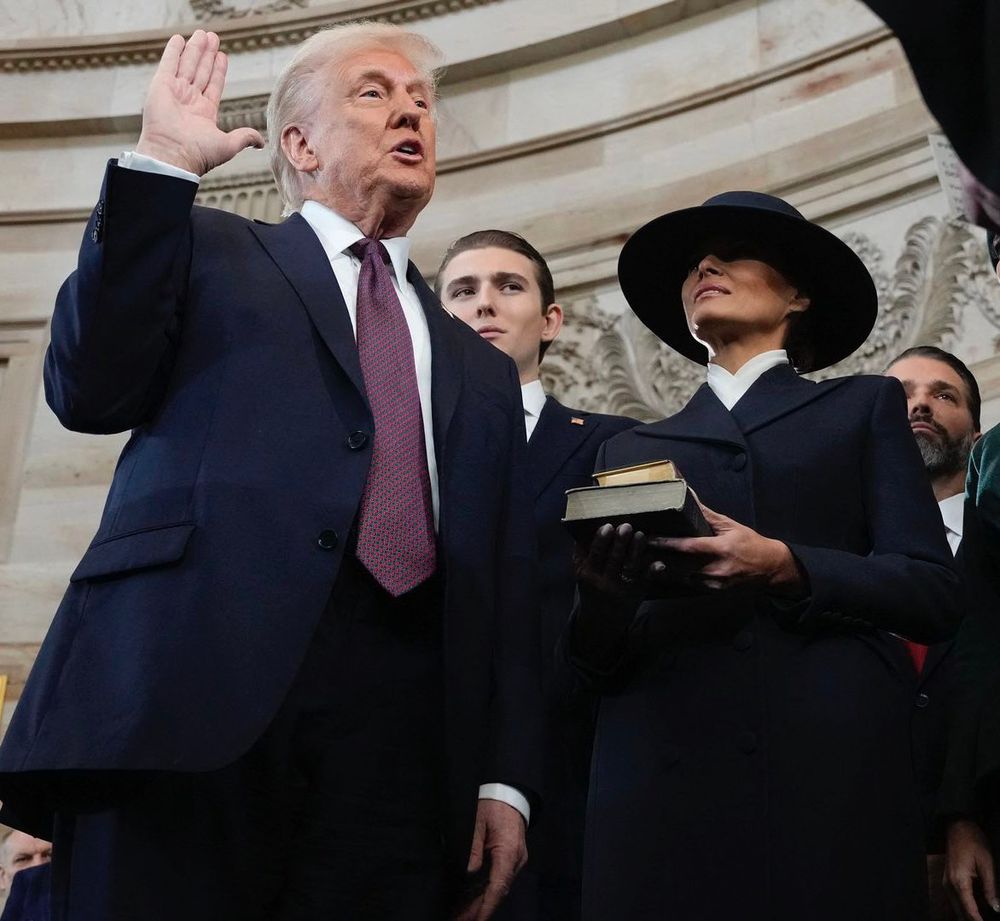 The image captures a moment during a ceremonial event, featuring a man with light-colored hair, dressed in a dark suit, raising his right hand in an oath-taking gesture. He appears to be speaking, with a serious expression. Beside him stands a woman wearing a wide-brimmed black hat and a dark coat, holding a book in her hands. A young man in a suit is positioned behind them, observing the proceedings. The setting appears to be an official indoor location with ornate architectural details in the background, suggesting a formal occasion.