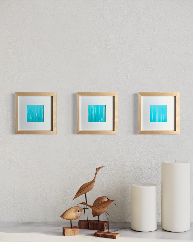 Three abstract prints of teal blue framed in maple are centered on a light colored wall. Below on a shelf are four wooden carved birds with two white pillar candles to the right. 