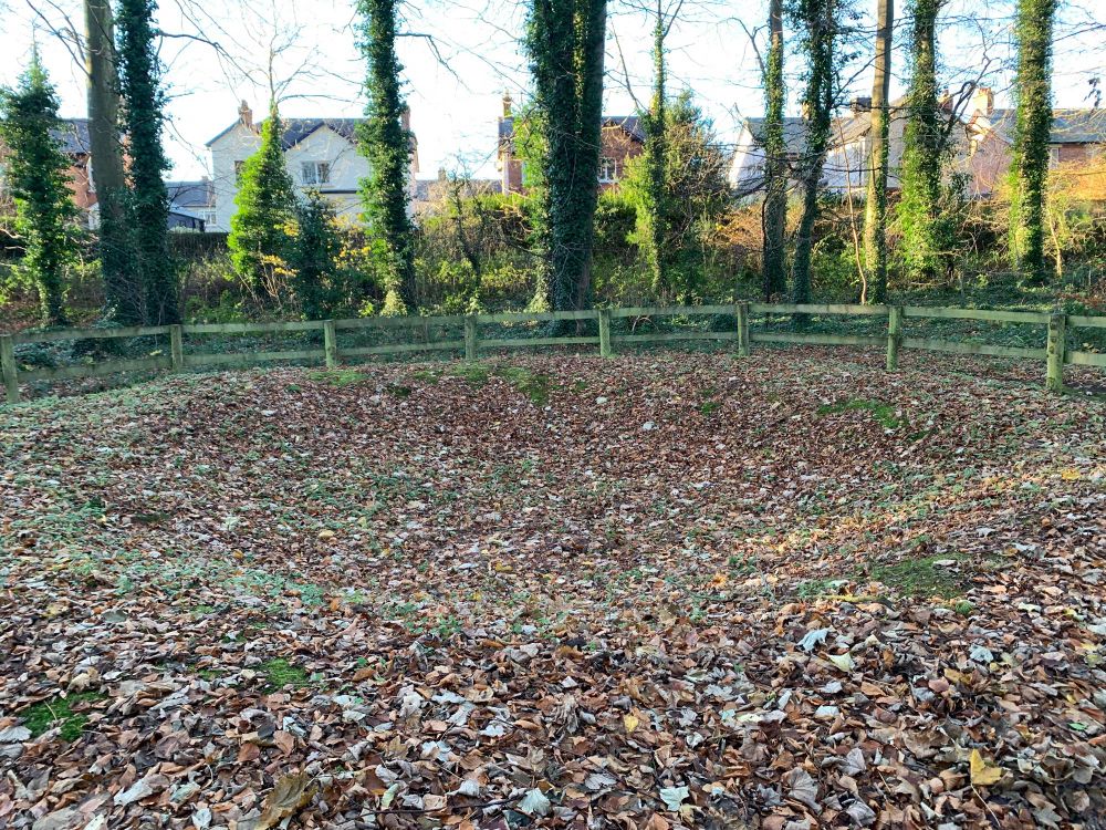 Photograph of bomb crater covered in fallen leaves and surrounded by wooden fencing.