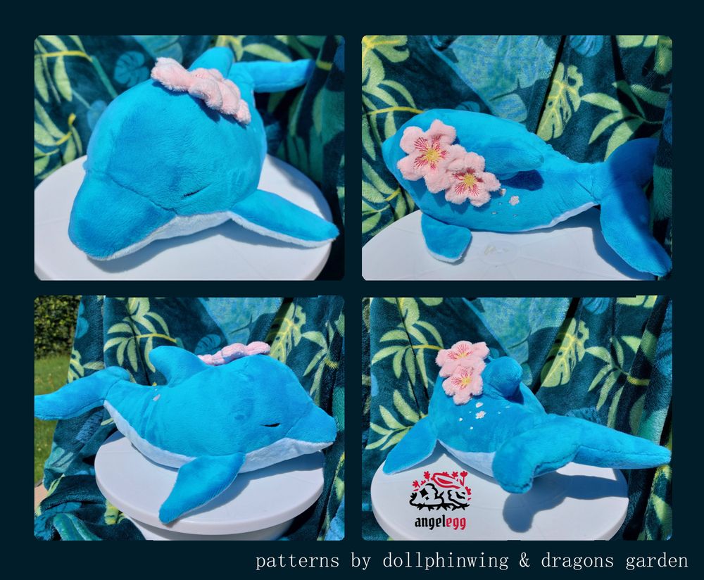 4 photos of varying angles of a handmade dolphin plushie. the dolphin has 2 cherry blossom plush sewn to the top near it's dorsal fin, with some cherry blossom petal embroidery going down it's back

at the bottom is written "patterns by dollphinwing & dragons garden"