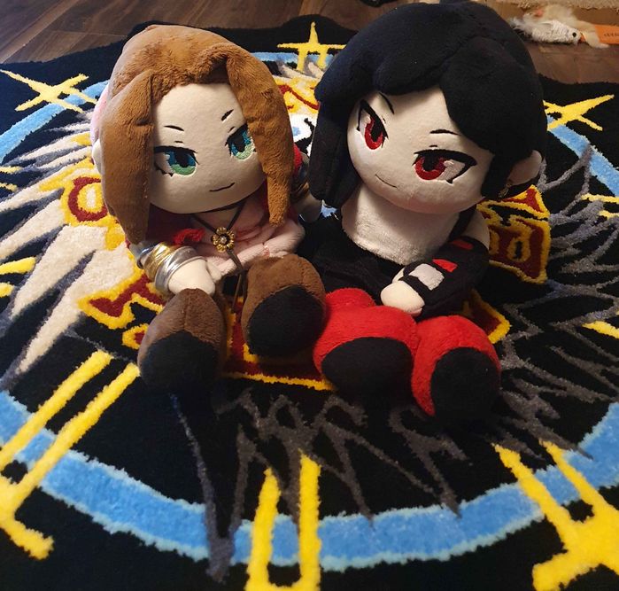 photo of 2 handmade plushies based on aerith and tifa from final fantasy VII sitting atop a handmade rug of the seventh heaven logo from the same game