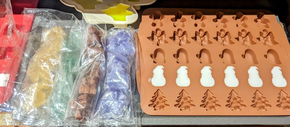 A silicone christmas mold with a row of snowman shaped soaps poured into it. Beside it are 4 plastic bags filled with different coloured soaps, all being prepared for Advent Calendars.