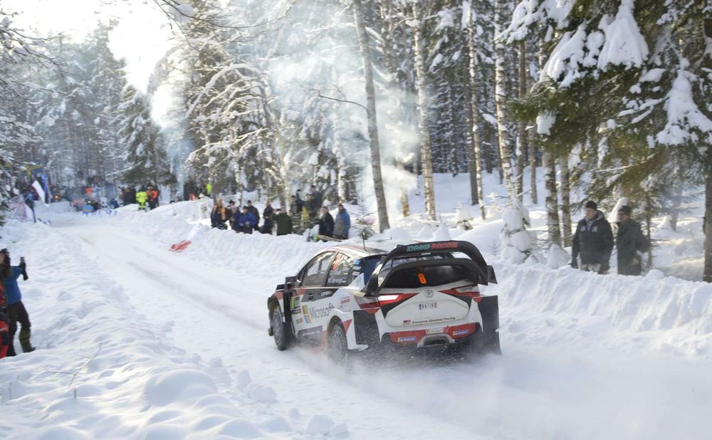 Rally car yeeting down a snowy forest track away from camera with spectators lining the snowbanks as it goes