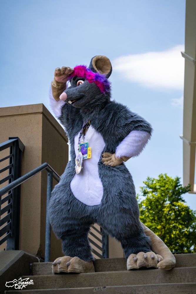 A female rat fursuit is standing on top of the stairs, looking to the side with one hand on forehead and the other on waist.