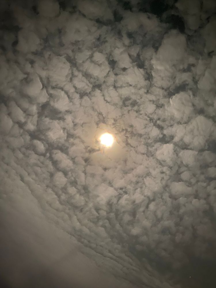 An upward shot of the night sky, from a grocery store parking lot. The bottom left corner has a faint glow from a distant and unseen lamp. The clouds mostly cover the sky with ‘cracks’ revealing the dark space beyond. The moon, though obscured by clouds, is bright and pierces through. 