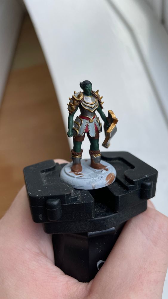 WIP of Kagra, the orc cleric of Lathander, a 3D printed miniature base coated in silver with gold trim and red and white detail. She has a silver shield with gold trim, green skin and black hair, and a bad attitude