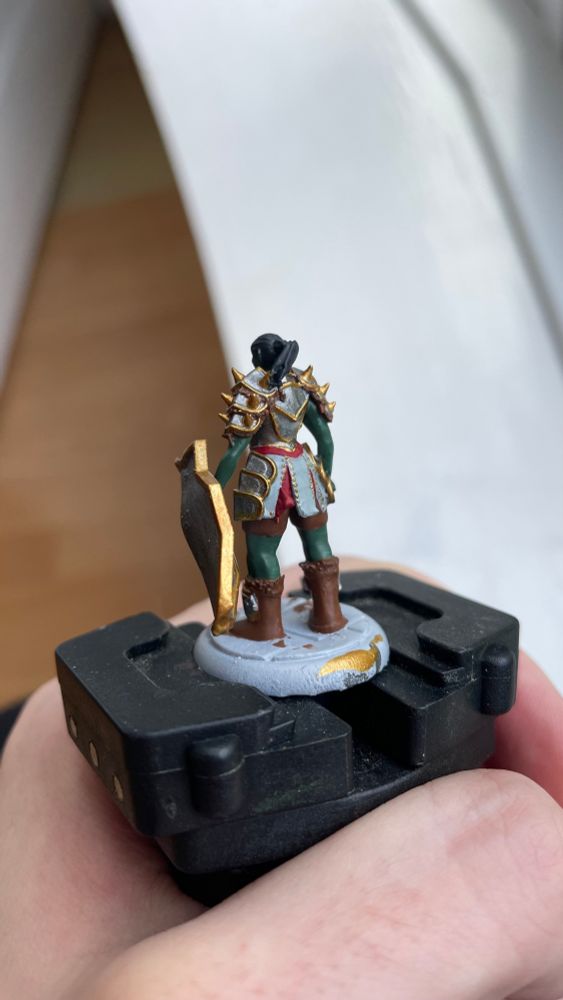 WIP of Kagra, the orc cleric of Lathander, a 3D printed miniature base coated in silver with gold trim and red and white detail. She has a silver shield with gold trim, green skin and black hair, and a bad attitude