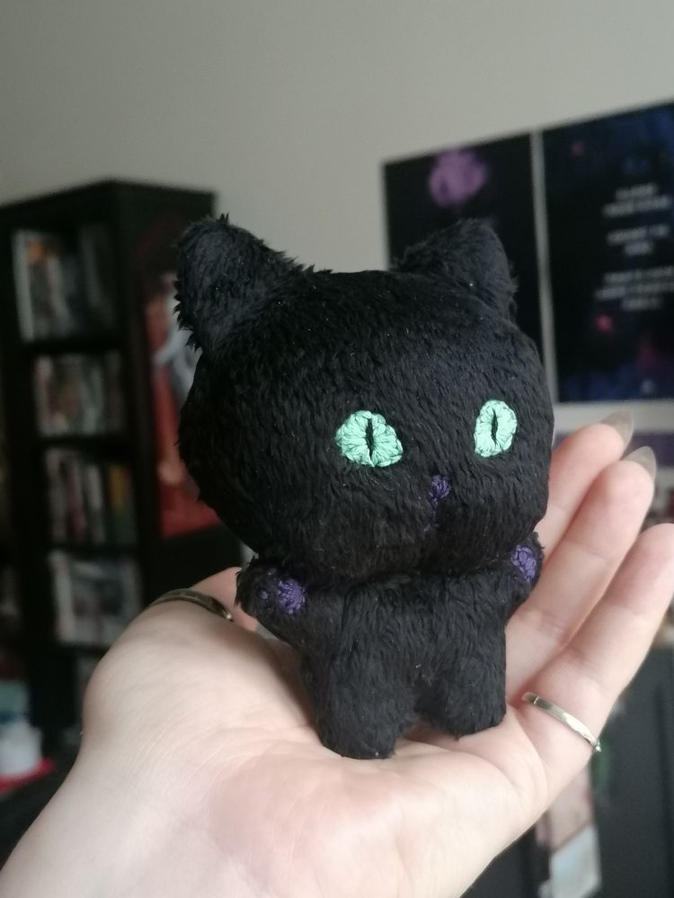 a small "chibi" plush doll of a black cat, held up in a hand, 3/4 view