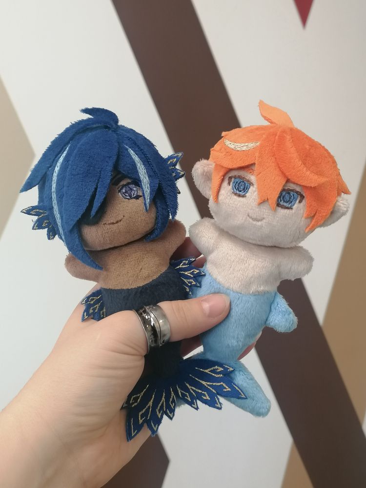 a photo of a hand holding two small plushies: a dark skinned merman with dark blue hair and tail with gold decorated fins, and a light skinned merman with ginger hair and a light blue tail