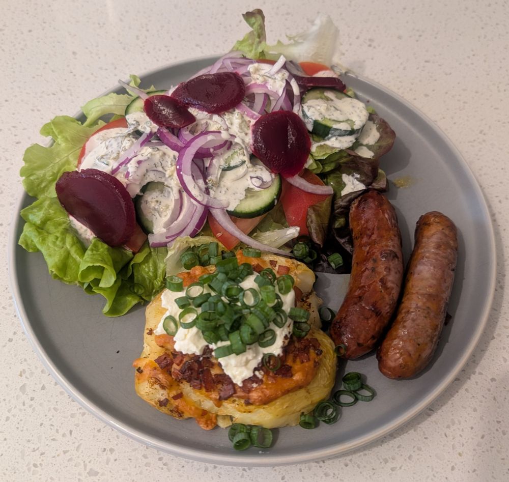 Salad consisting of sliced beetroot, sliced Lebanese cucumber, sliced tomato, sliced red onion, lettuce with ranch dill dressing on it. Smashed baked potato with melted cheese, Portuguese spices, diced crispy bacon, sour cream, chopped shallots and 2 beef sausages in a grey plate