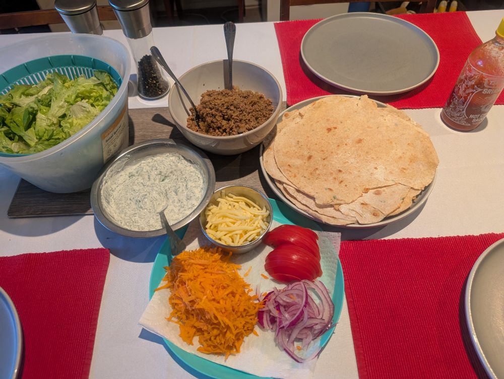 Cooked wholemeal wraps on a plate, 3 empty grey plates, aqua plate with grated carrot, grated cheese in a small bowl, sliced tomato, sliced red onion on paper towel wholemeal wrap cooking , bowl of cooked mince in a bowl,, bowl of mint yoghurt, bowl of lettuce on a table
