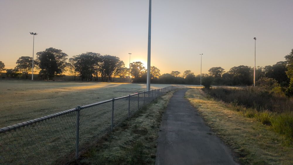 Football field with dew on the grass, concrete path, trees in the background with the sun slowly coming up
