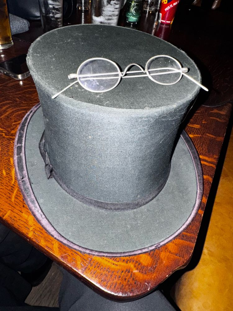 Picture of a top hat on a pub table, on the hat are glasses worn by Ralph Ineson as Wilhelm Sievers in Robert Eggers’ film Nosferatu 
