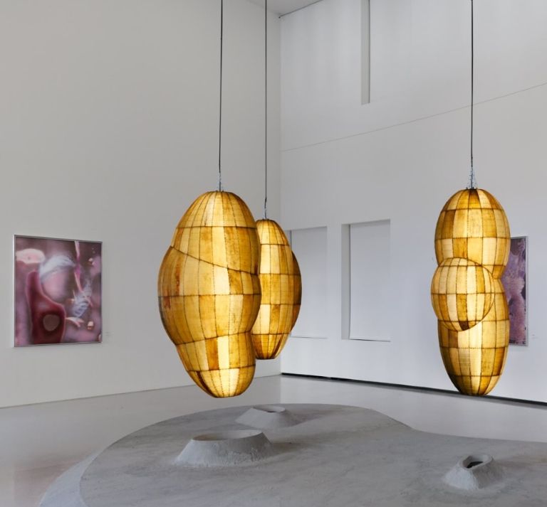 Gold pod like sculptures suspended from the ceiling of a gallery