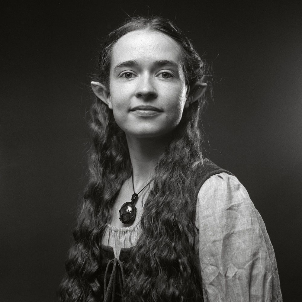 Black and white film photo of Nicole looking at the camera, in a hobbit dress a stone Necklace and curly hair, half up, revealing pointed ears. 