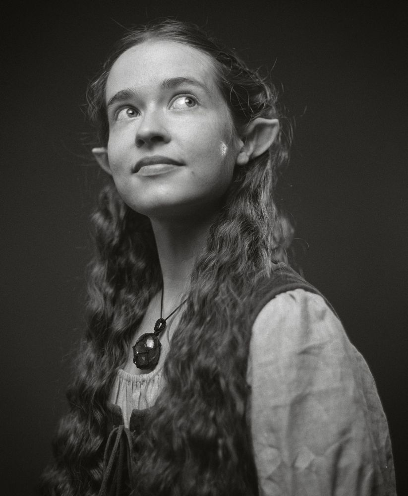 Black and white film photo of Nicole looking up towards the right corner, in a hobbit dress a stone Necklace and curly hair, half up, revealing pointed ears. 