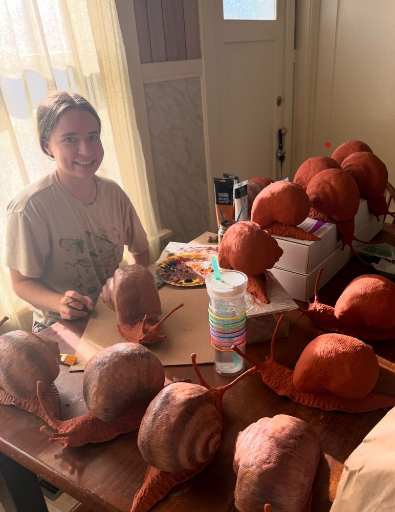 Nicole sits at a table with some paint. The table is covered with sculptures of snails, climbing all over. Nicole has a smile but internally she is slightly concerned because she realizes that she is a mother now to so many snails.