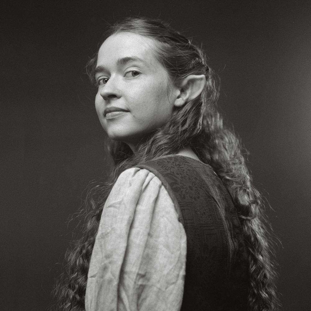 Black and white film photo of Nicole looking at the camera over her shoulder, in a hobbit dress a stone Necklace and curly hair, half up, revealing pointed ears. 