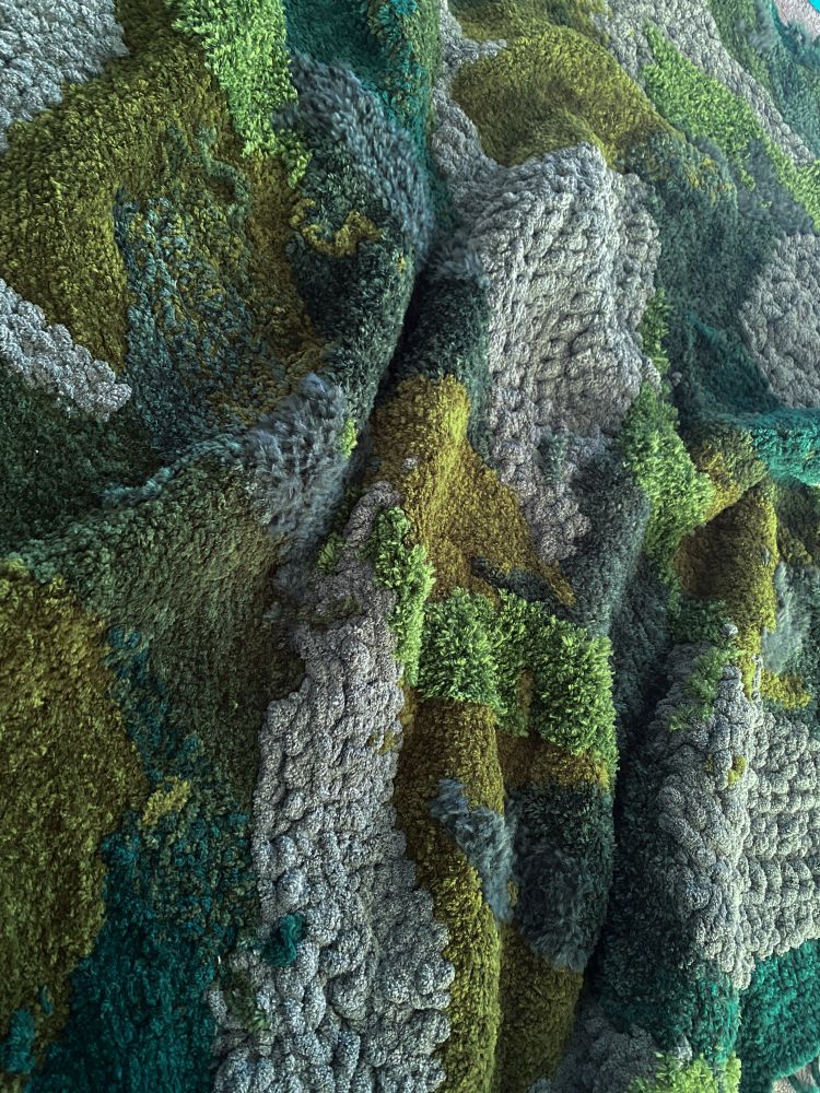 A crocheted blanket that is a patchwork of a variety of green fuzzy yarns that resemble Moss. 