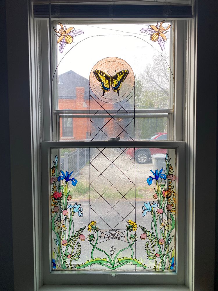 A window with craft paint to give the illusion of stain glass. The bottom has a floral design and the top has a swallowtail and stag beetles. 