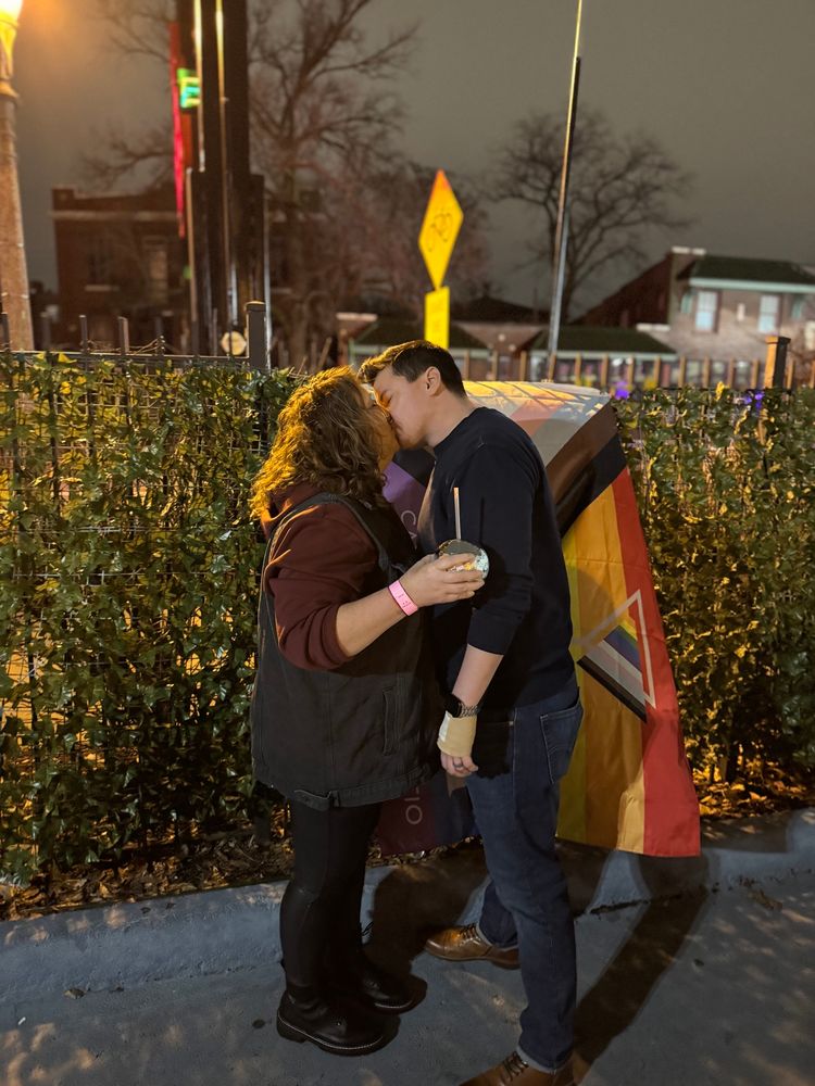 Poster and spouse sharing a kiss in front of pride flag