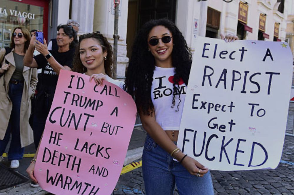 Two Romans holding signs 1- reads: I’d call Trump a cunt, but he lacks depth and warmth. 2- reads: elect a rapist expect to get fucked