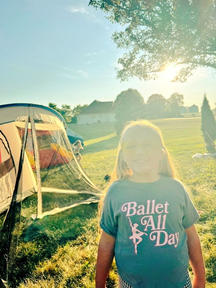 Little blonde girl wearing a "Ballet All Day" t-shirt, standing next to a tent in a grassy field. She looks glowing because she was photographed with the sun behind her 