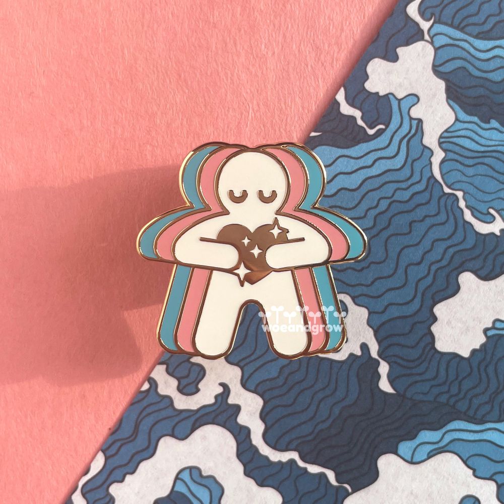 Photo of my Trans Love enamel pin of a humanoid figure with a calm expression holding a sparkling gold heart to their chest with colors radiating out from them to form the trans pride flag