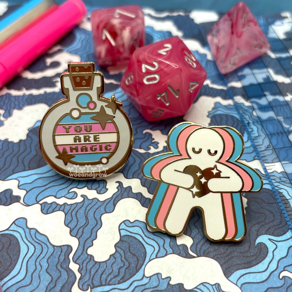 Closeup photo of my You Are Magic and Trans Love enamel pins on a blue wave patterned background surrounded by pink dice and some matching desk supplies
