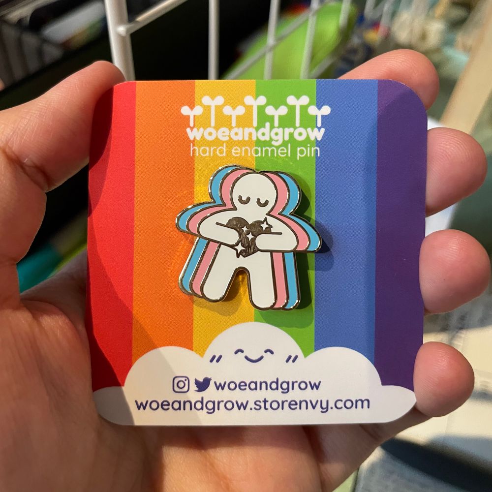 Photo of my Trans Love enamel pin on rainbow backing card held in hand