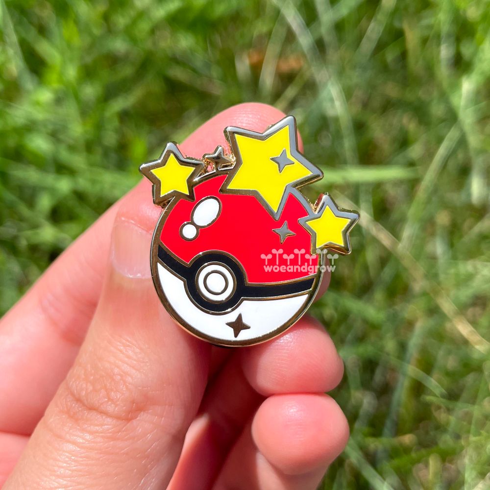 Closeup photo of my enamel pin of a Poke Ball with stars as if it just successfully caught a Pokemon, in my hand with grass out of focus in the background 