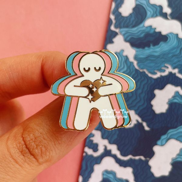 Closeup photo of my Trans Love enamel pin of a figure with eyes closed in a calm expression holding a sparkling heart to their chest and colors radiating out from them to form the transgender pride flag