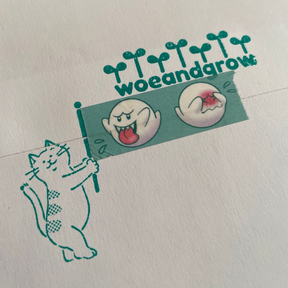 Photo of a stamp of a cat holding a flag pole in its front paws with washi tape as the flag. Next to it is my woeandgrow logo stamp both in green ink on a white envelope.