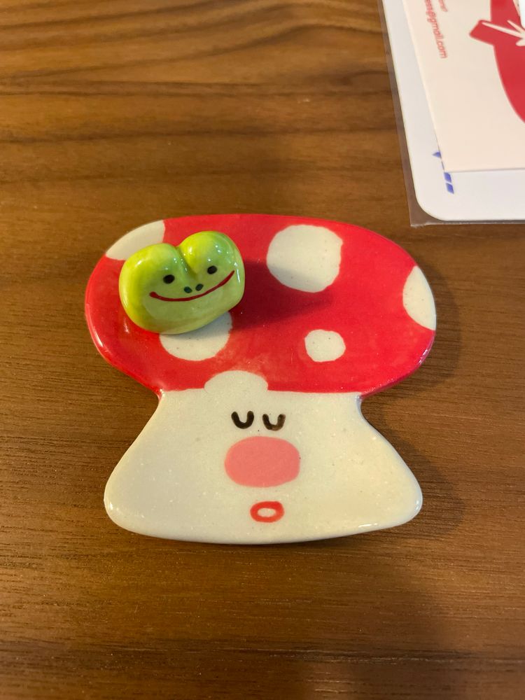 Photo of a small ceramic mushroom dish with a calm expression and a small smiling ceramic frog head figure sitting on top.