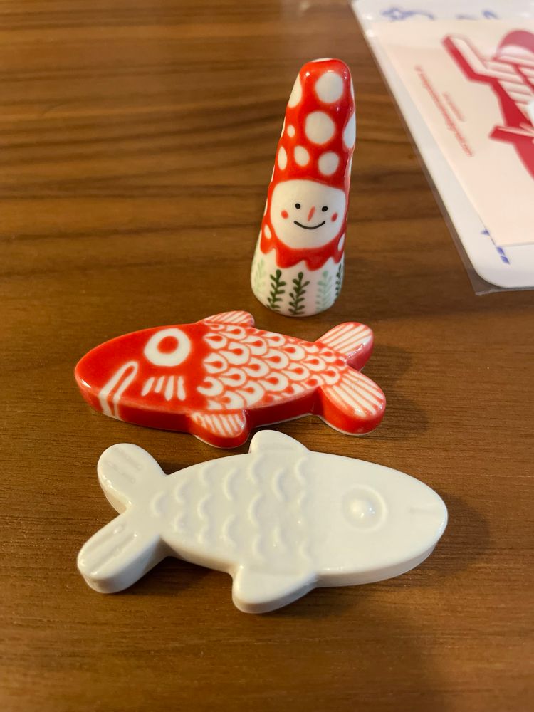 Photo of two ceramic fish chopstick rests, one red and white and one all white, and a ring holder figure with a smiling face, a red and white spotted hat, and green leafy details