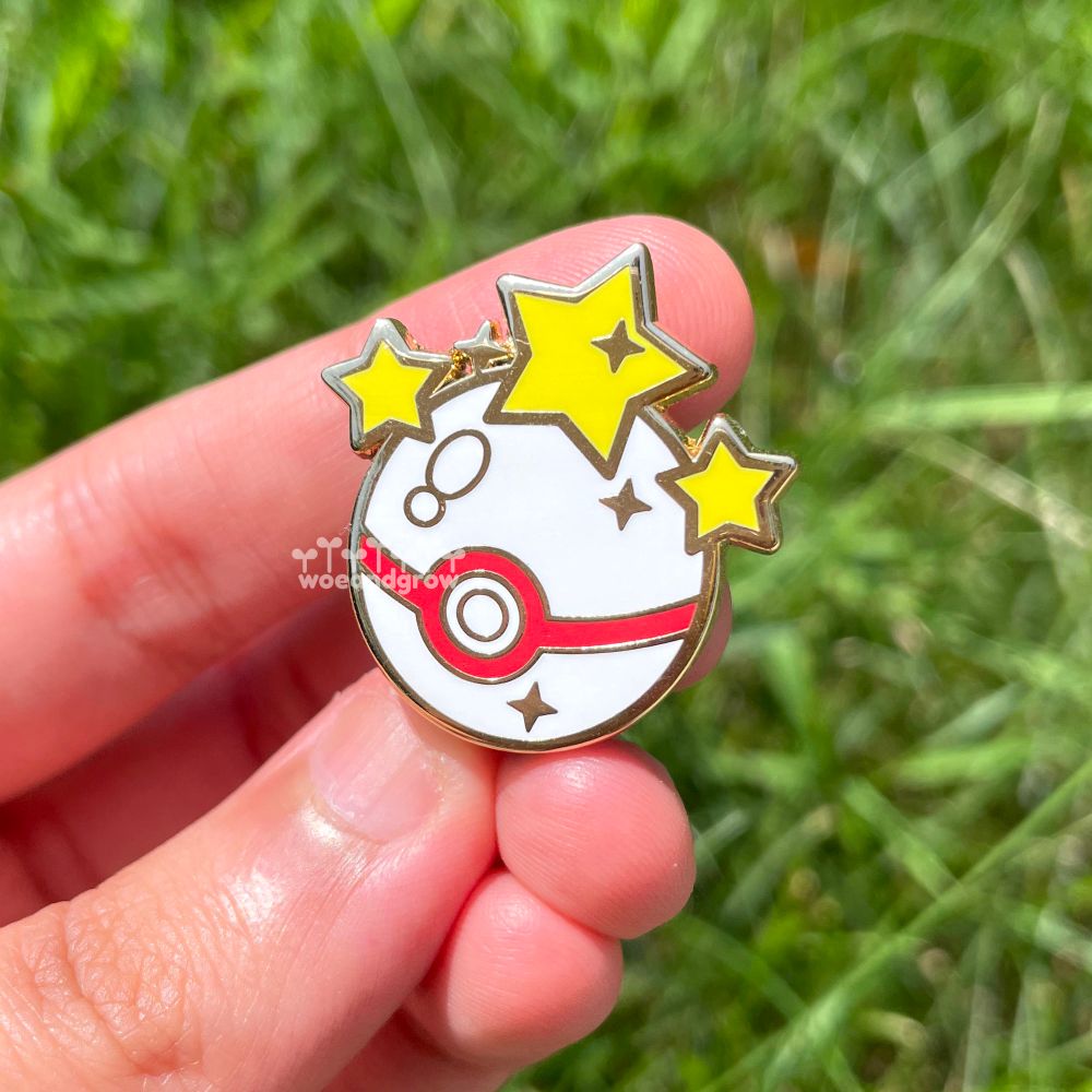 Closeup photo of my enamel pin of a Premier Ball with stars as if it just successfully caught a Pokemon, in my hand with grass out of focus in the background 