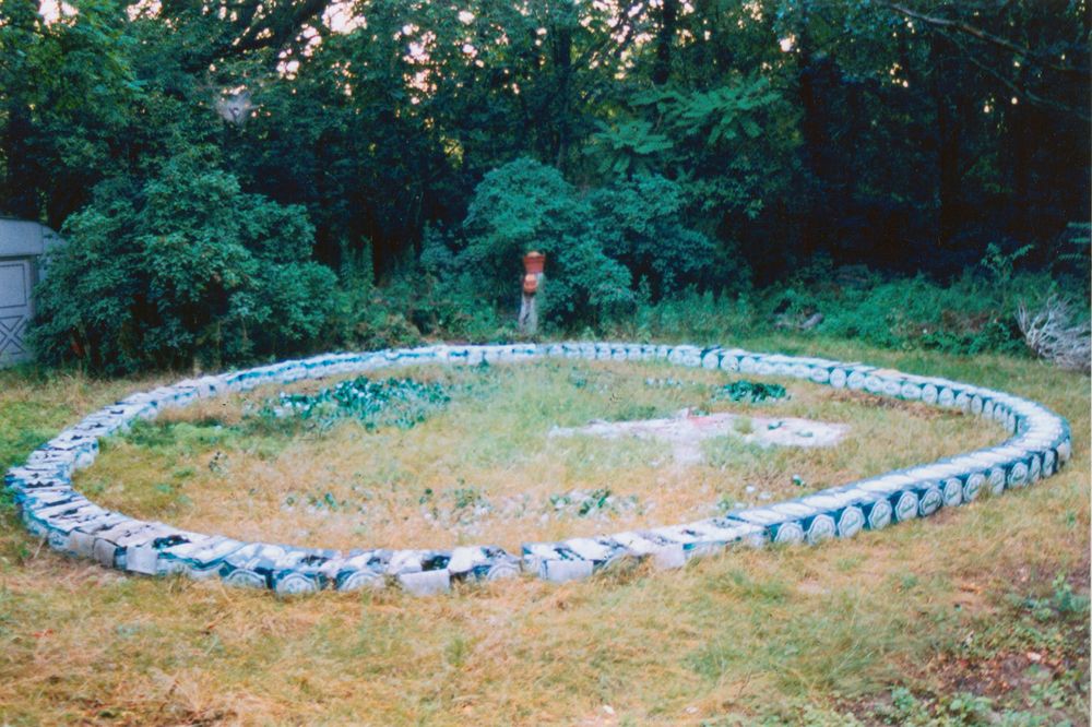 Boxes that look to be cardboard boxes once held cans or bottles are arranged on grass in a circle near the edge of some woods.