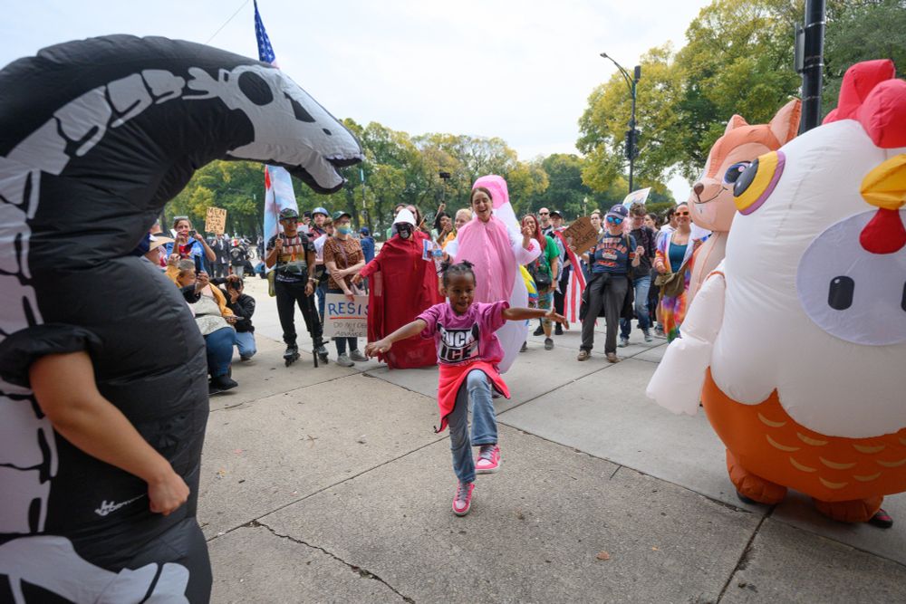 A kid wearing a "Fuck ICE" shirt dances among inflatable animals as people cheer behind them.