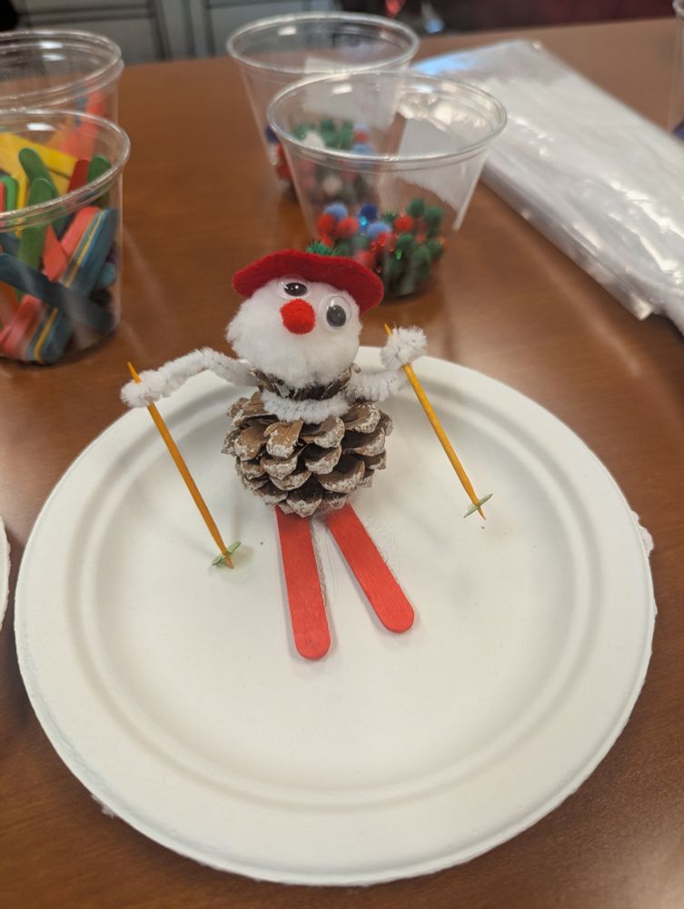 A little snowman made of a pine cone, a cotton ball on top for a head  some pipe cleaners for arms and a couple of ice cream sticks for skis. This one has a red felt hat and red skis. 