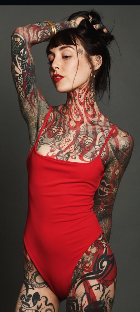 Very thin woman wearing a bright red swimsuit. Her body is converted in overlapping tattoos: she has a regular set of line at tattoos in dark ink, and a second set of thick wavy lines, almost like stylistic Arabic script, overlaid over the other layer.