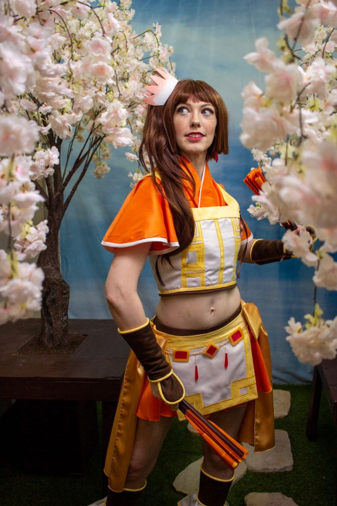 A photo of me in my Tsuruno Yui cosplay from Magia ReCord. It's a white, orange, and yellow magical girl outfit that is vaguely reminiscent of a waitress uniform because Tsuruno's family owns a canonically mediocre Chinese restaurant. Behind me are some faux sakura trees and a blue backdrop.