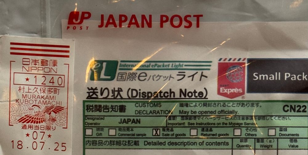 This is a close-up photo of a shipping label from a package sent via Japan Post. 

Key details visible in the image:
- **Japan Post** is printed in bold red letters at the top.
- There is a white and green section labeled "**International ePacket Light**" and "Dispatch Note."
- The label includes a "**Small Packet**" and “Express” icon.
- On the left, there is a red postage stamp with Japanese writing. It says:
    - "NIPPON" (Japan)
    - "1240" (which may be the postage amount)
    - “村上久保多町 MURAKAMI KUBOTAMACHI” (the post office location)
    - A decorative bird illustration
    - The date: "18.07.25" (July 25, 2018)
- The customs declaration section lists “JAPAN” as the origin and checkboxes for various package types like "Commercial sample" and "Sale of goods," none of which appear selected.

The label is primarily in Japanese and English. The overall look is official, structured, and busy with text, boxes, and small print, typical for international parcels.
